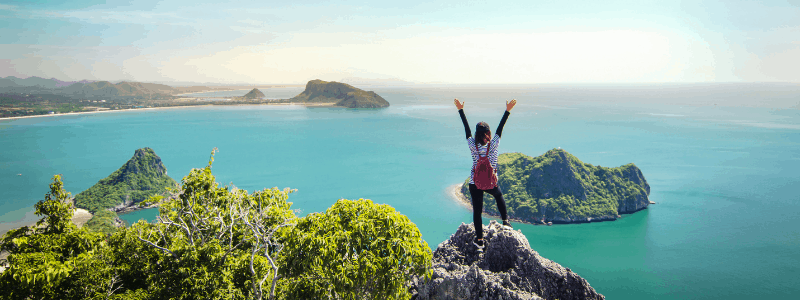 Photo of a person stood on top of a mountain with the arms raised in the air, looking out over the ocean.