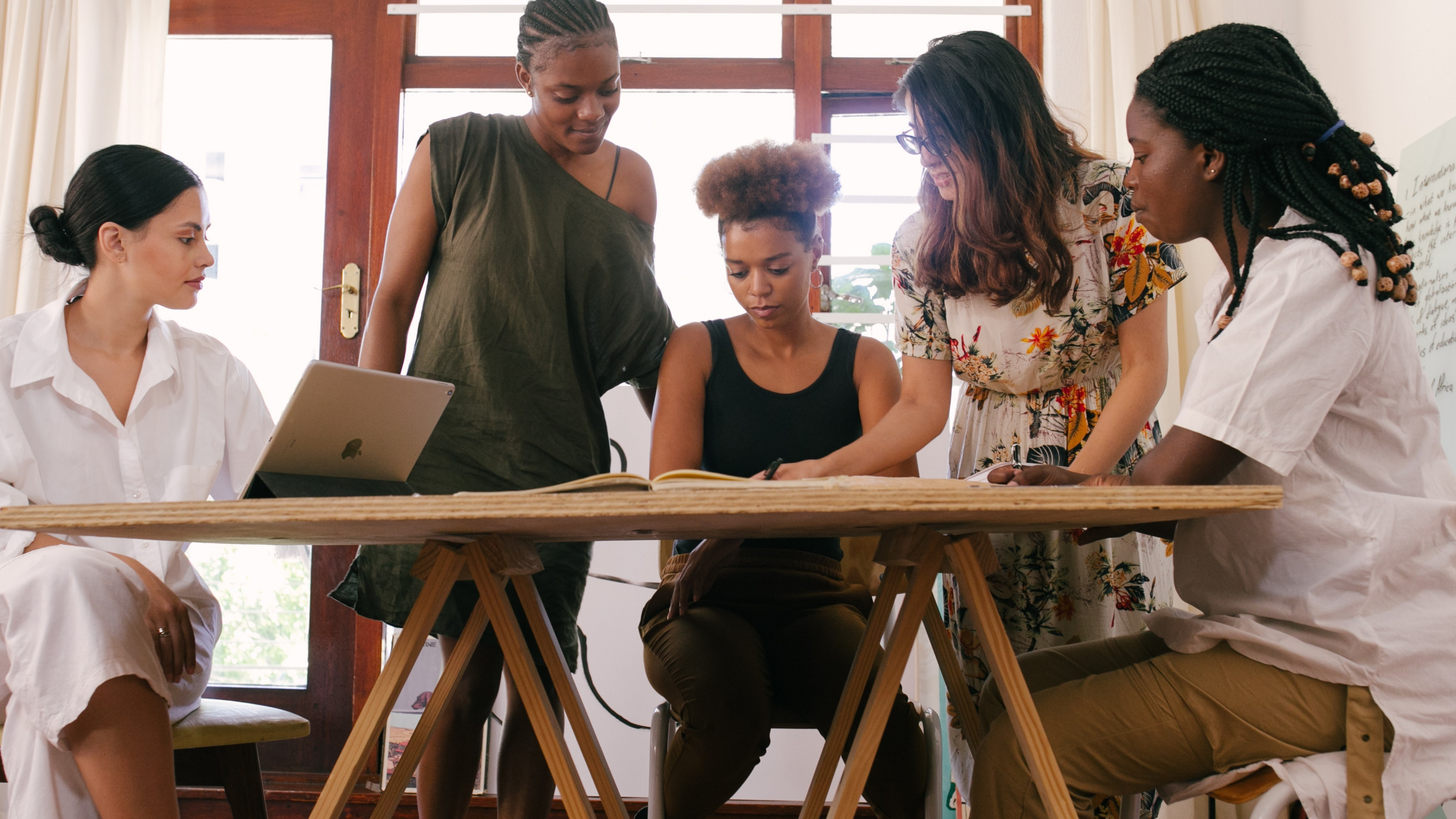 Five women sitting and standing around a desk looking at each others work.